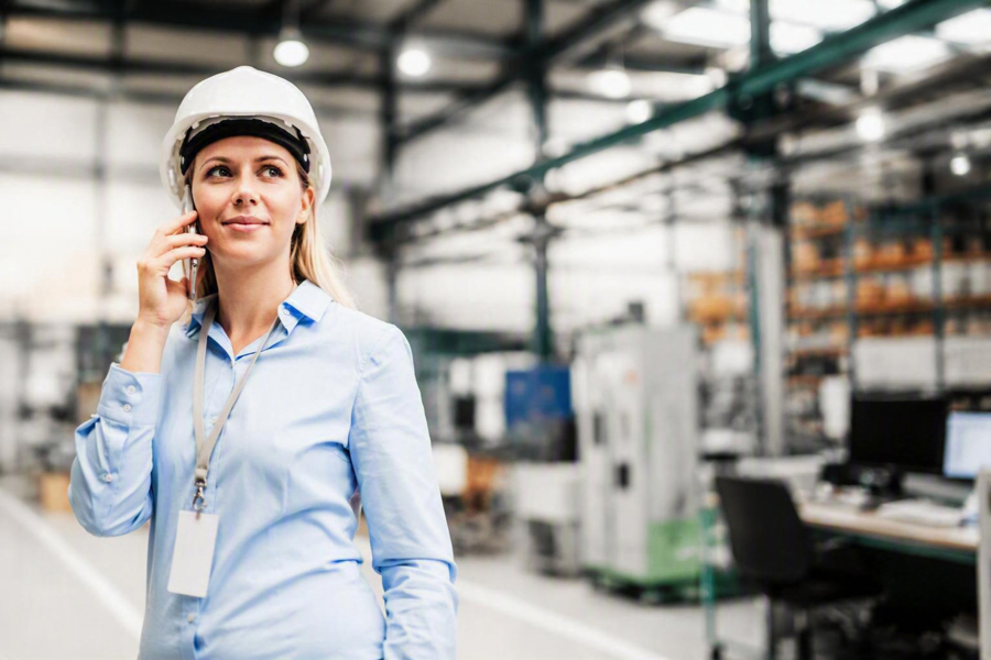 Person med skyddshjälm använder mobiltelefon i industrimiljö med produktionsutrustning i bakgrunden