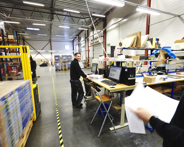 Medarbetare på Electro Elco arbetar i lager- och produktionsmiljö med orderhantering, logistikflöden och kvalitetskontroll vid arbetsstation nära lager och trucktrafik