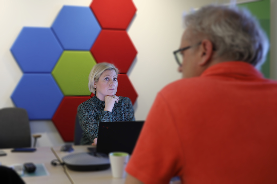 Person sitter vid mötesbord med laptop medan kollega lyssnar, i kontorsmiljö med färgglada hexagoner på väggen i bakgrunden