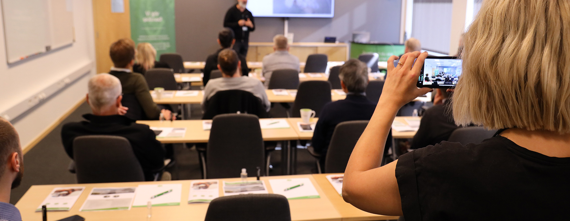 Seminarie i konferensrum där en person presenterar framför en skärm medan deltagare sitter vid bord, och en person i förgrunden håller upp en mobil för att ta bild