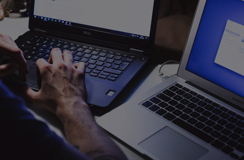 Närbild av händer som skriver på en laptop, med en andra laptop bredvid i en mörk arbetsmiljö och skärmar som lyser upp tangenterna