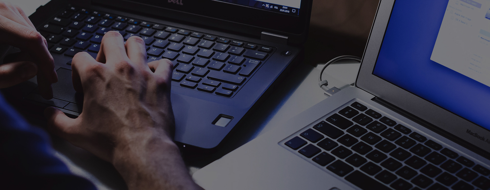 Närbild av händer som skriver på en laptop, med en andra laptop bredvid i en mörk arbetsmiljö och skärmar som lyser upp tangenterna