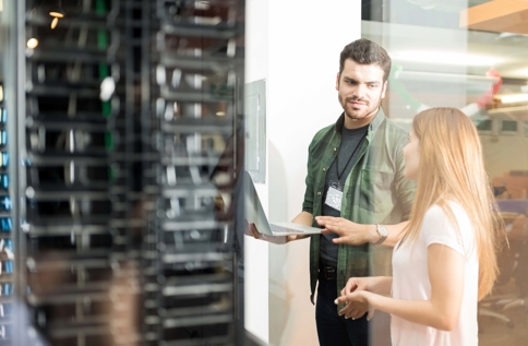 Två personer står vid serverrack i modern IT‑miljö där den ena visar information på laptop, vilket illustrerar serverdrift som tjänst, samarbete, övervakning och hantering av IT‑infrastruktur i datacenter