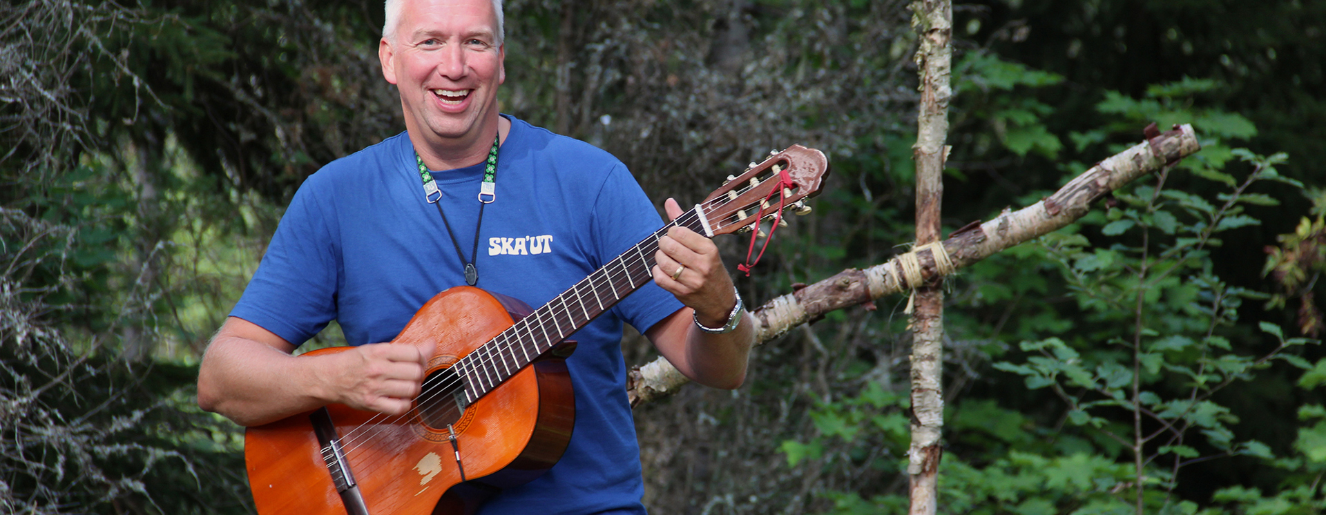 Stefan Sjöberg står utomhus i skogsmiljö och spelar gitarr, med träd och grönska i bakgrunden