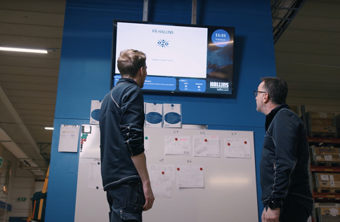 Medarbetare följer produktionsinformation på digital informationsskärm i industrimiljö med fokus på kommunikation, planering och verksamhetsstyrning