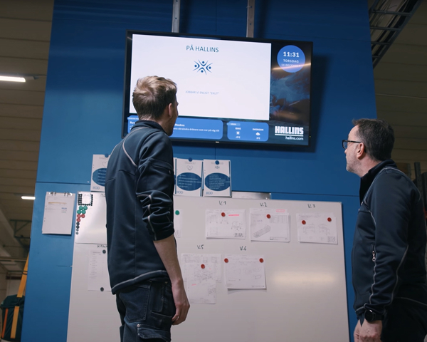 Medarbetare följer produktionsinformation på digital informationsskärm i industrimiljö med fokus på kommunikation, planering och verksamhetsstyrning