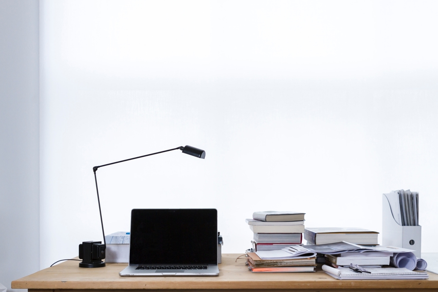 Arbetsbord med laptop, skrivbordslampa och staplar av böcker och dokument mot en ljus, enkel bakgrund
