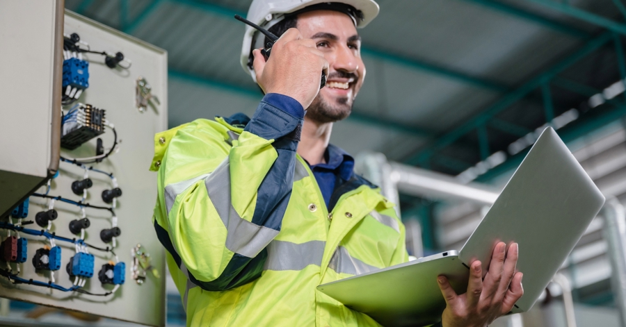 Medarbetare I Varselkläder Använder Laptop Och Kommunicerar Via Headset I Modern Industrimiljö Med Produktionsutrustning I Bakgrunden