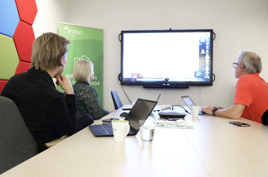 Tre personer sitter vid konferensbord i digitalt mötesrum och arbetar med laptop och stor skärm för presentation, samarbete och videomöten i modern kontorsmiljö