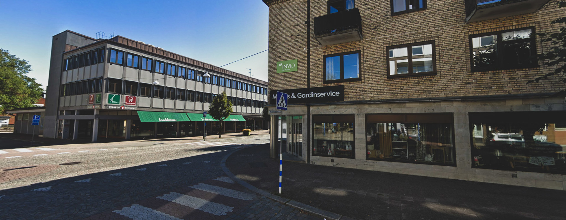 Gatubild i Vetlanda med äldre byggnader i tegel och puts, där en grön INVID‑skylt syns på en fasad, samt butikslokaler och korsning i förgrunden under klar himmel