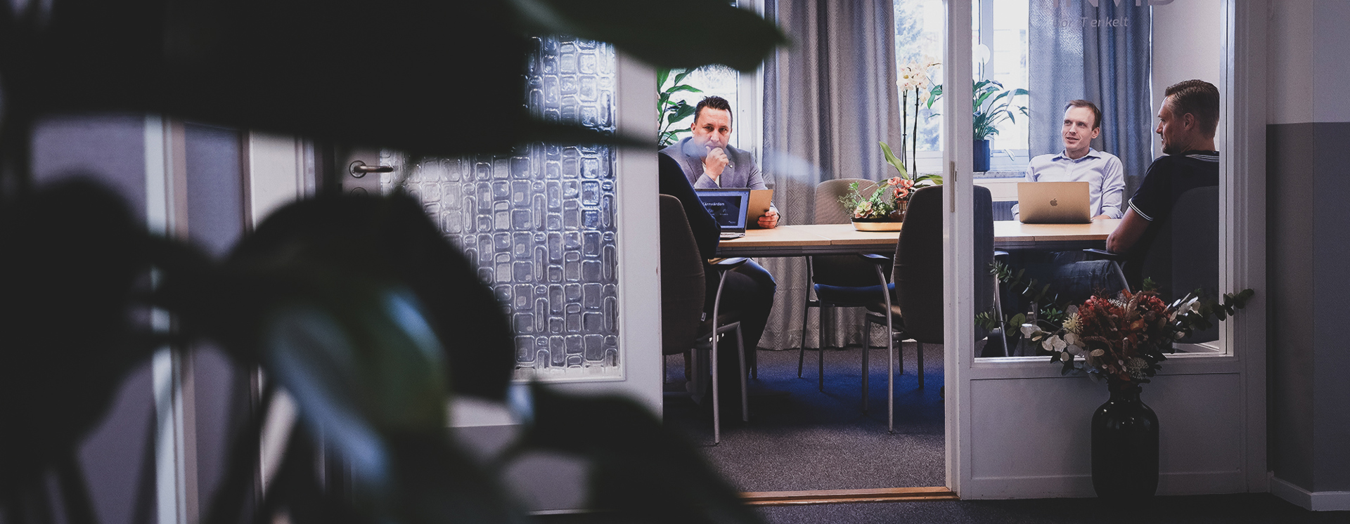 Fyra personer sitter runt ett bord i ett mötesrum med laptops, sett genom en glasvägg med växter i förgrunden