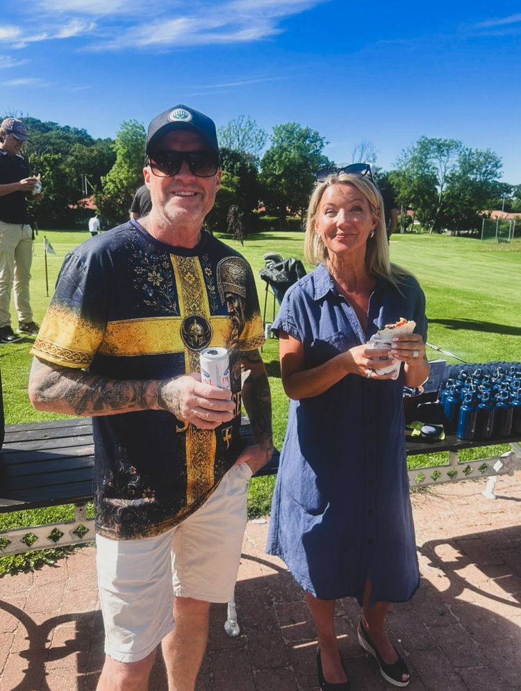 Deltagare vid golfevent står på terrassen och äter lunch med golfbanan i bakgrunden under en solig sommardag