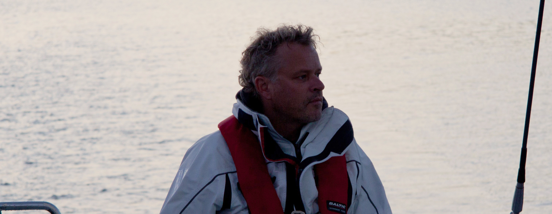 Joakim Hermansson står vid ratten på en segelbåt och styr, med flytväst och jacka, omgiven av öppet vatten i bakgrunden