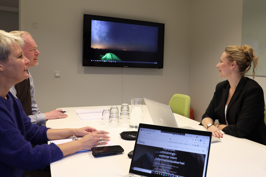Tre personer sitter runt konferensbord i mötesrum och diskuterar framför väggmonterad skärm med presentation, tillsammans med laptop, anteckningar och dokument för samarbete och digitalt möte