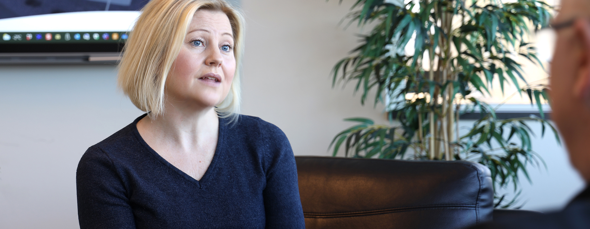 Maria Burman sitter i en fåtölj i ett kontor och samtalar med en annan person utanför bild, med en stor grön växt och skärm på väggen i bakgrunden
