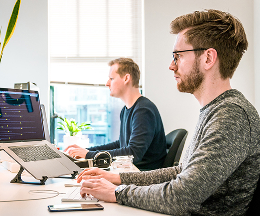 Två personer arbetar vid skrivbord med laptop och extern skärm i modern kontorsmiljö, vilket visar servicedesk, IT‑support och övervakning av system och ärenden i verksamheten