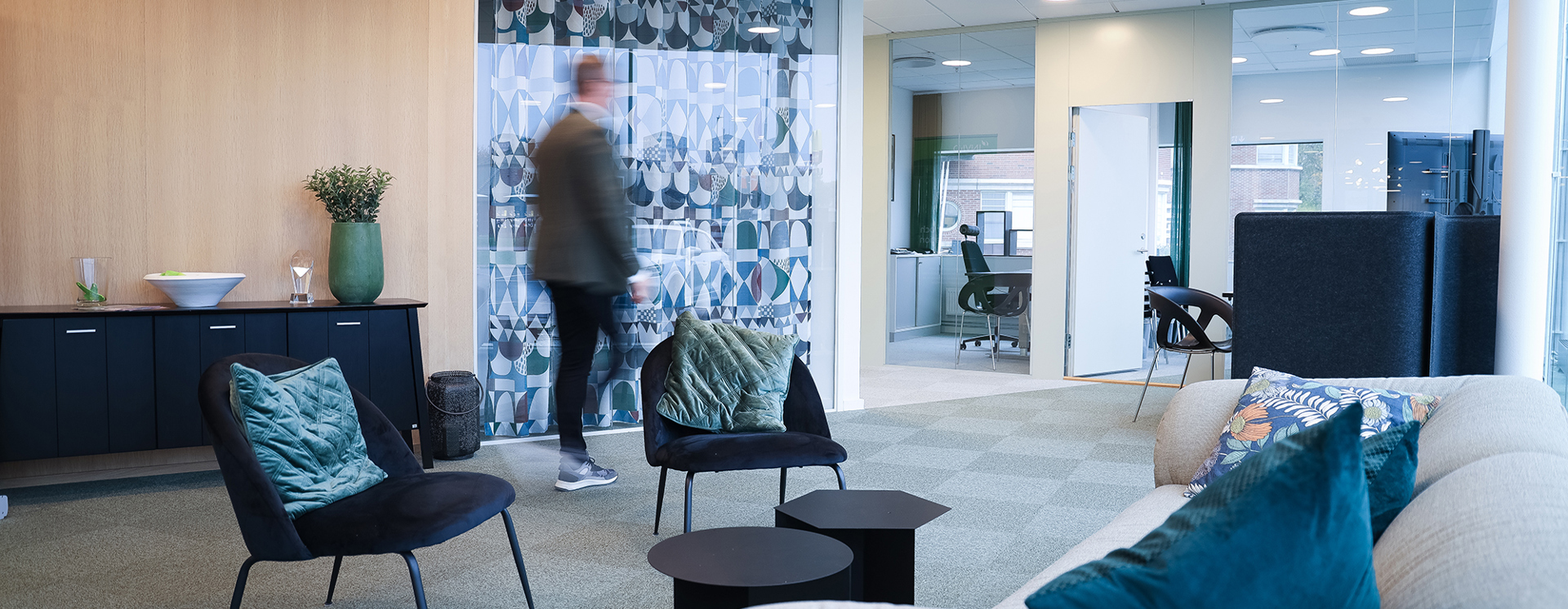 Modern loungeyta i kontor med fåtöljer, soffa och små bord, där en person går förbi framför en glasvägg och öppna arbetsrum