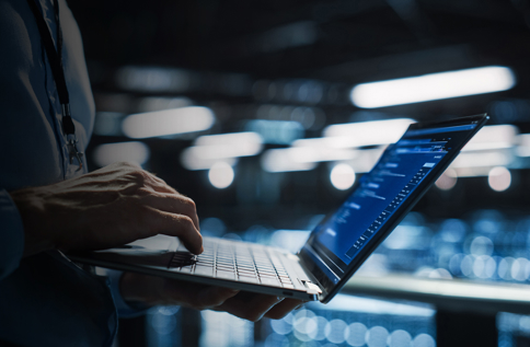 Person arbetar på laptop i serverhall med bakgrund av datacenter och nätverksutrustning, vilket visar övervakning, drift och underhåll av IT‑miljö och affärssystem i realtid
