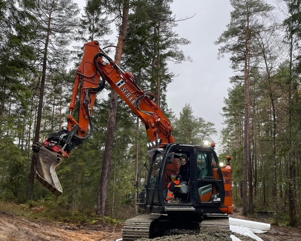 Bandgrävare arbetar med mark- och anläggningsarbete i skogsmiljö där väg eller infrastruktur byggs med hjälp av tung entreprenadutrustning