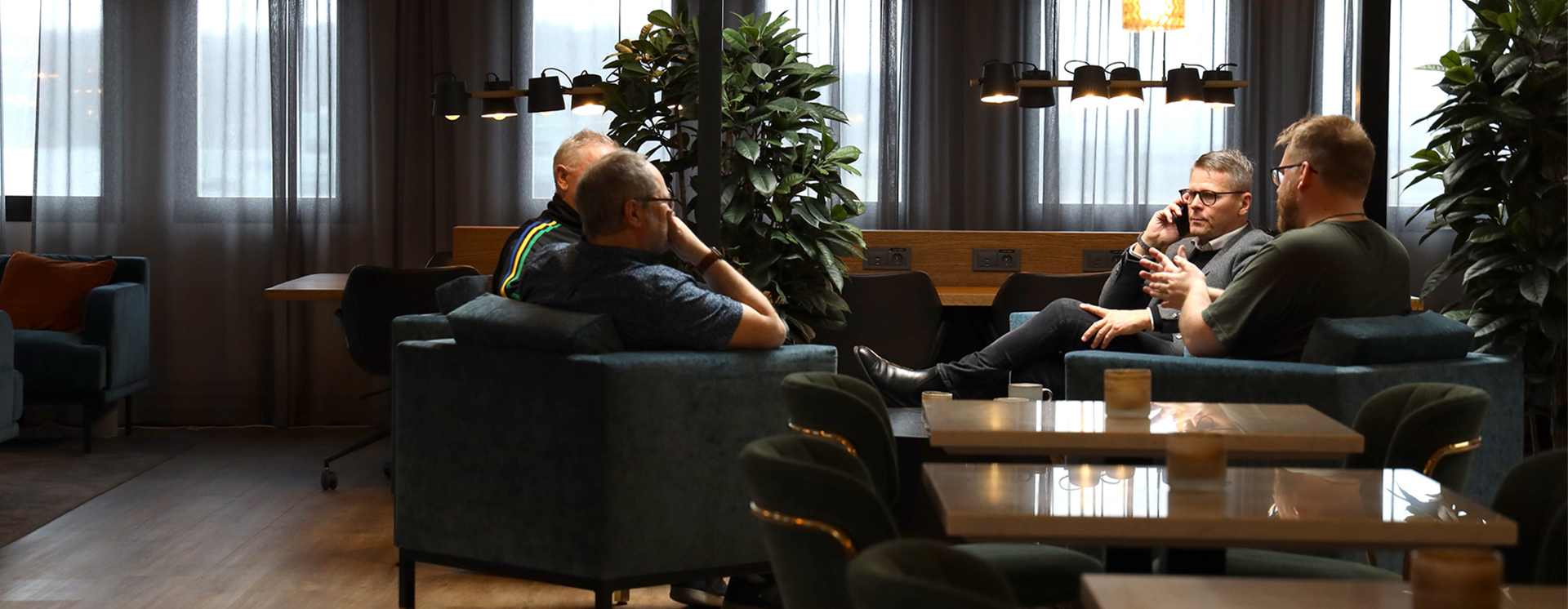 Fyra personer sitter i en loungeyta i ett kontor och samtalar i soffor och fåtöljer, med bord, växter och arbetsplatser i bakgrunden
