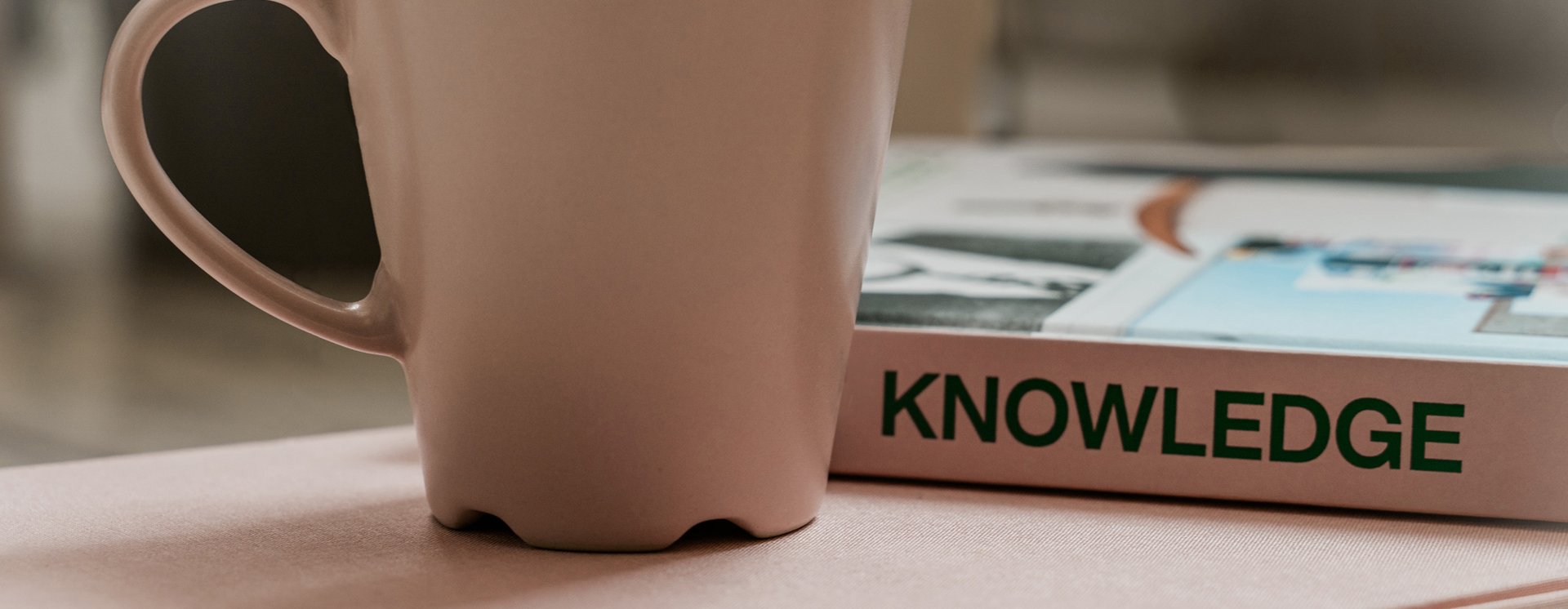 Närbild av en ljus kopp på ett bord bredvid en bok med texten “KNOWLEDGE” på ryggen, i en mjukt upplyst inomhusmiljö