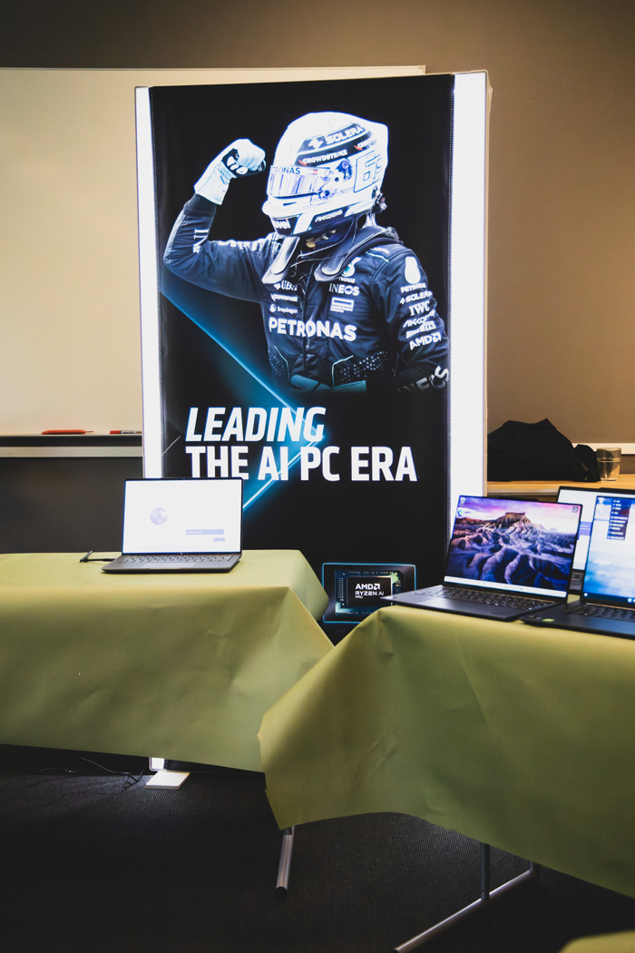 Teknikutställning med laptops uppställda på bord framför skylt med texten “Leading the AI PC Era” och bild på racerförare.