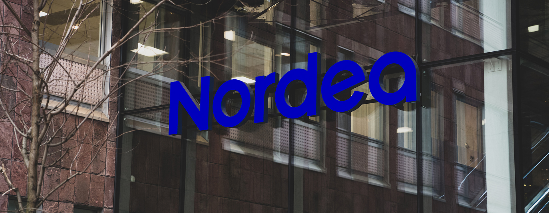 Entré med glasfasad där den blå Nordea-logotypen syns på byggnaden, med reflektioner av omgivande hus i glaset