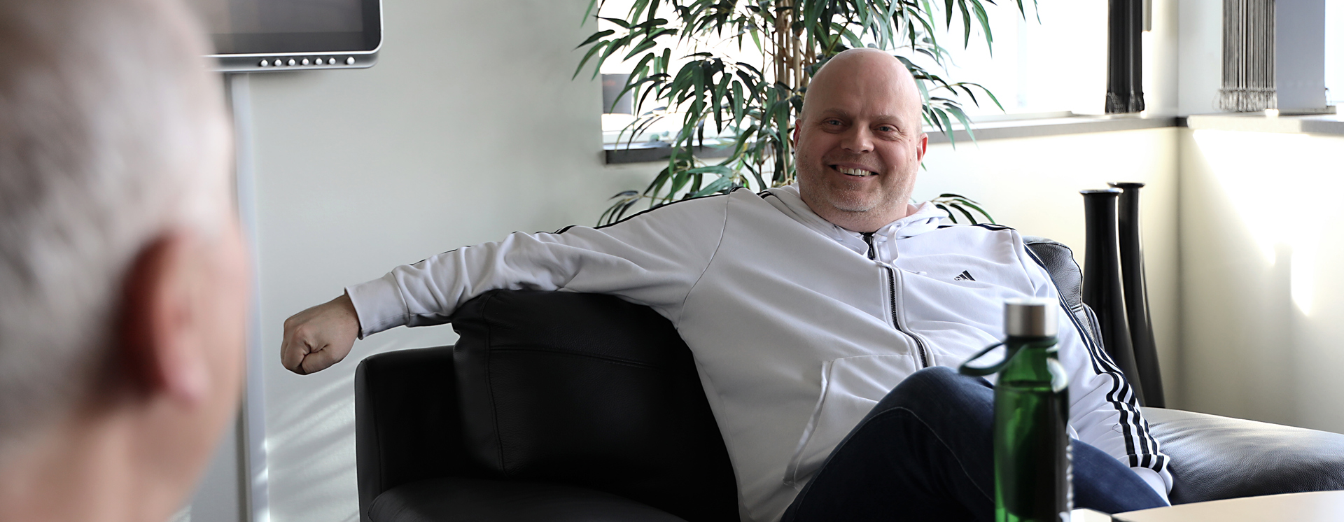 Tobias Hansson sitter i en fåtölj i ett kontor vid ett runt bord med vattenflaska och mobil, med en stor grön växt och fönster i bakgrunden samt en annan person i förgrunden