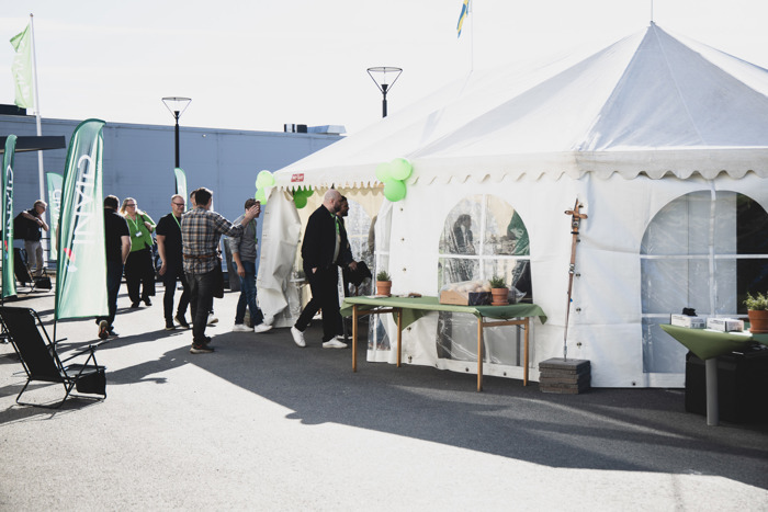 Personer minglar utomhus vid ett tält dekorerat med gröna ballonger under ett företagsevent, med INVID-flaggor och bord uppställda intill.