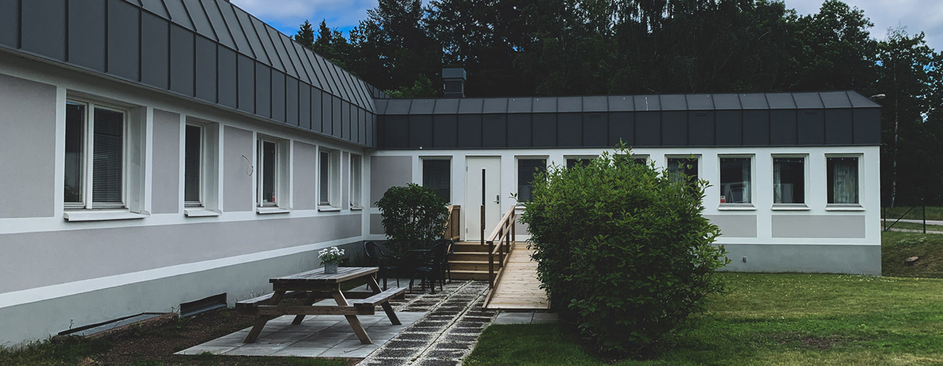 Enplansbyggnad i Tranås med flera fönster och mörkt tak, sedd från en gräsmatta med gångstig, uteplats, bänk och buskar i förgrunden, samt träd och molnig himmel i bakgrunden