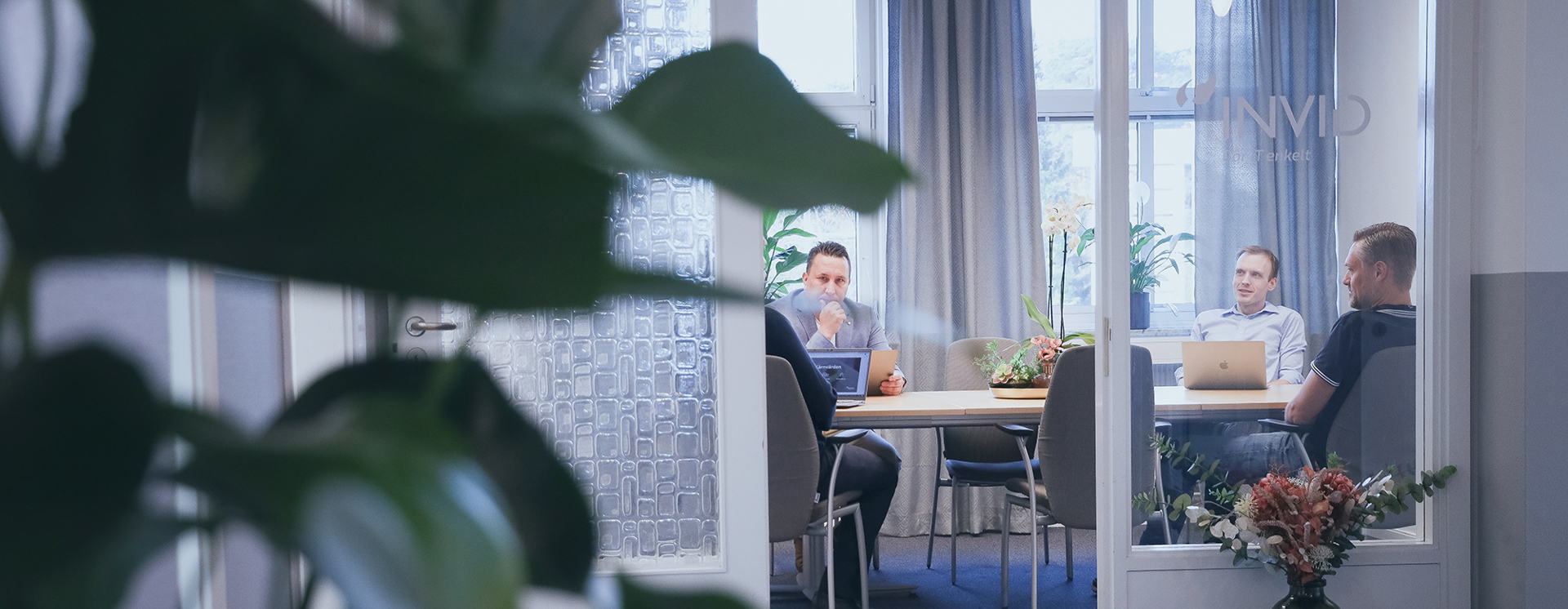 Fyra personer sitter runt ett bord i ett mötesrum och arbetar vid laptops, sett genom en glasdörr med växter i förgrunden