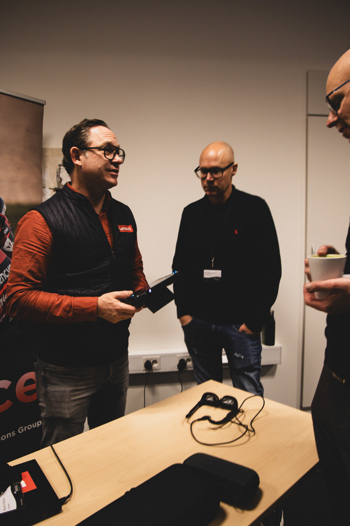 Tre personer står vid ett bord i ett konferensrum och samtalar, där en håller i en enhet, med VR‑glasögon på bordet och Lenovo-rollup i bakgrunden