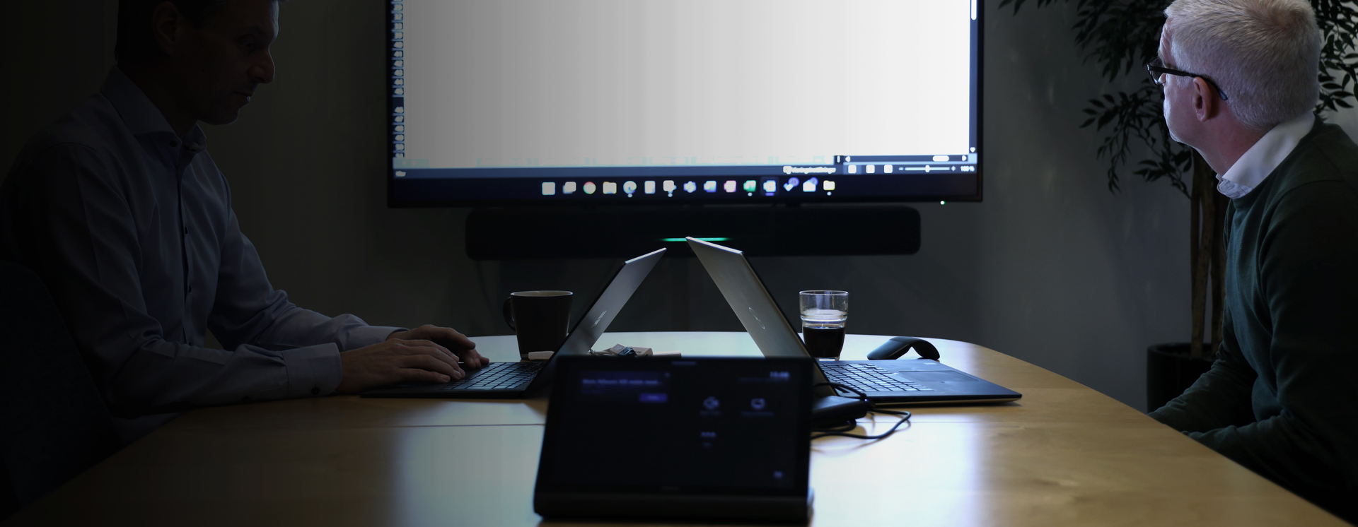 Två personer sitter vid konferensbord i digitalt mötesrum och använder laptop tillsammans med stor skärm för videomöte, presentation och samarbete i modern arbetsmiljö