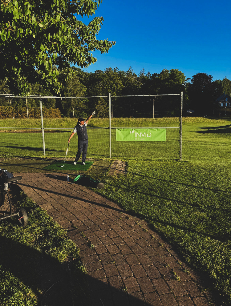 Golfspelare tränar slag vid driving range under företagsgolf med INVID-banner och grönskande golfbana i bakgrunden
