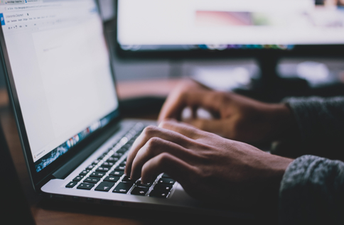 Händer som skriver på bärbar dator framför flera skärmar i kontorsmiljö med fokus på digitalt arbete och IT-tjänster