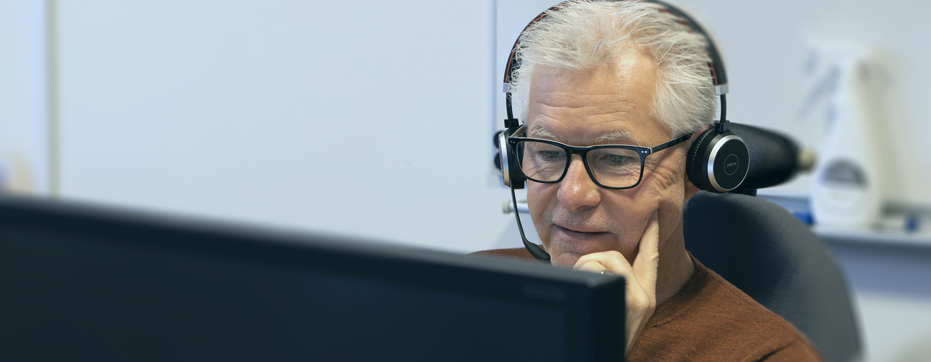 Person med headset sitter vid en dator och arbetar i en kontorsmiljö, sedd bakom en skärm i förgrunden