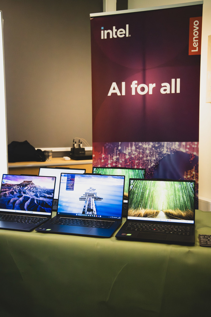 Laptops uppställda för demo på bord vid teknikutställning, framför roll‑up med texten “Intel – AI for all”.
