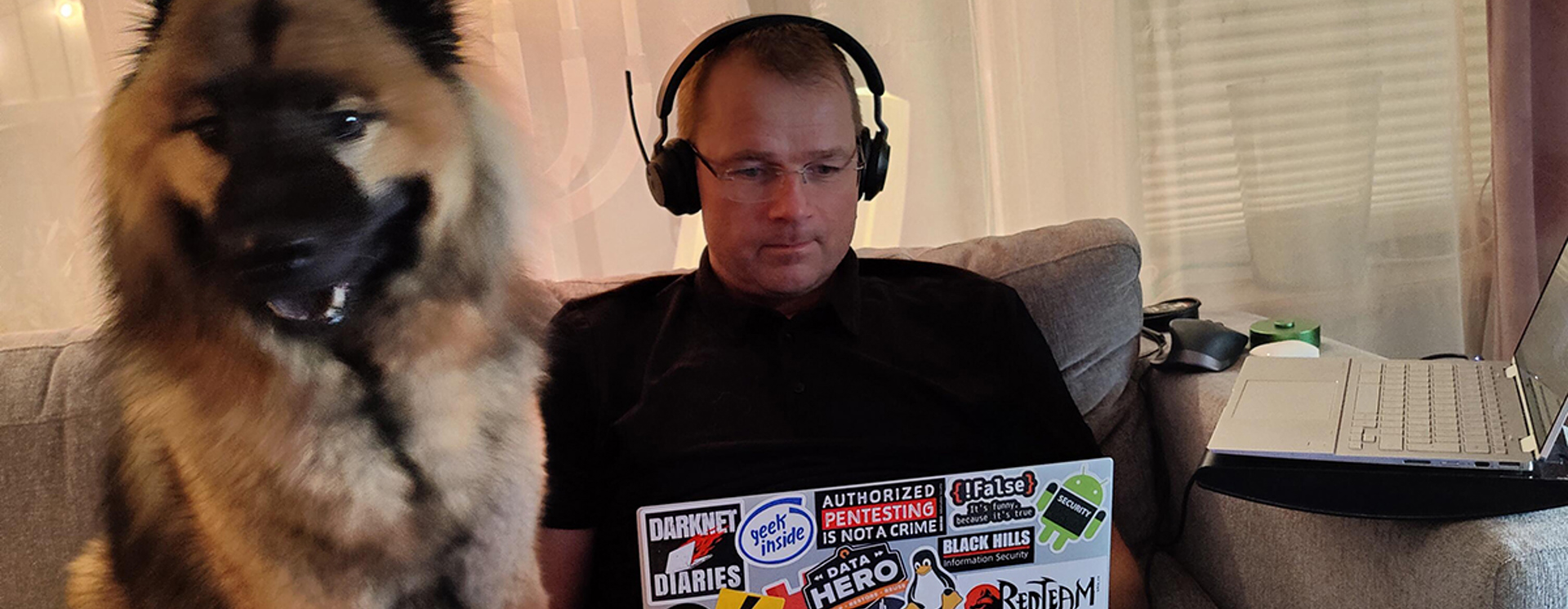 Roger Bergling sitter i en soffa och använder en laptop med klistermärken, med hörlurar på, medan en stor hund sitter bredvid honom i soffan