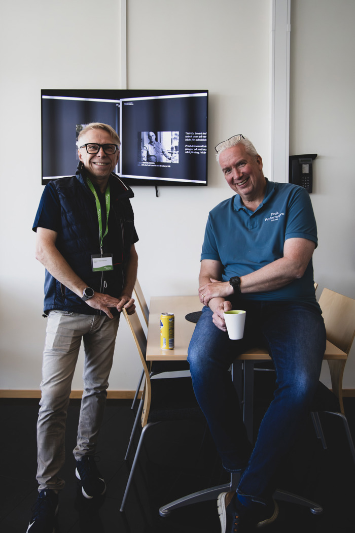 Två deltagare pausar och samtalar vid ett bord inomhus under event, med presentationsskärm i bakgrunden.